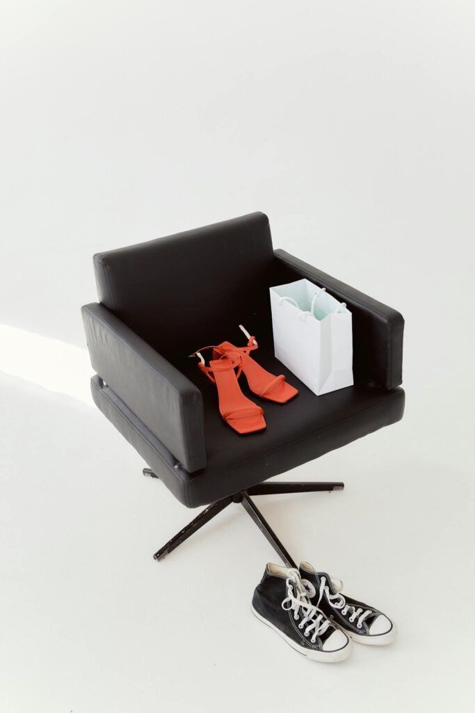 A swivel chair holds red high-heeled sandals and a shopping bag with sneakers below on a white background.